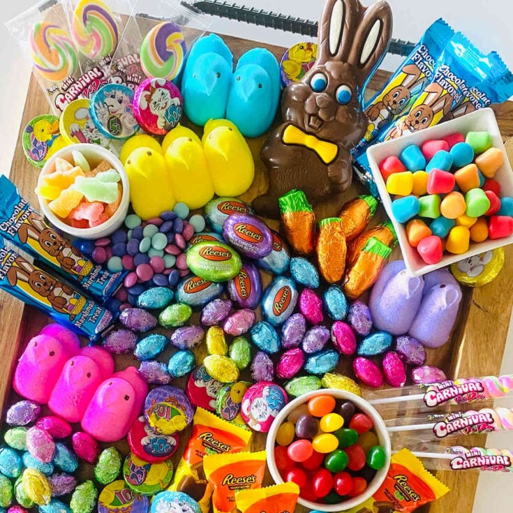 Marshmallow peeps, chocolate eggs, bright colored jelly beans, egg shaped Reeses candies and pastel colored lollipops on a wooden serving tray.