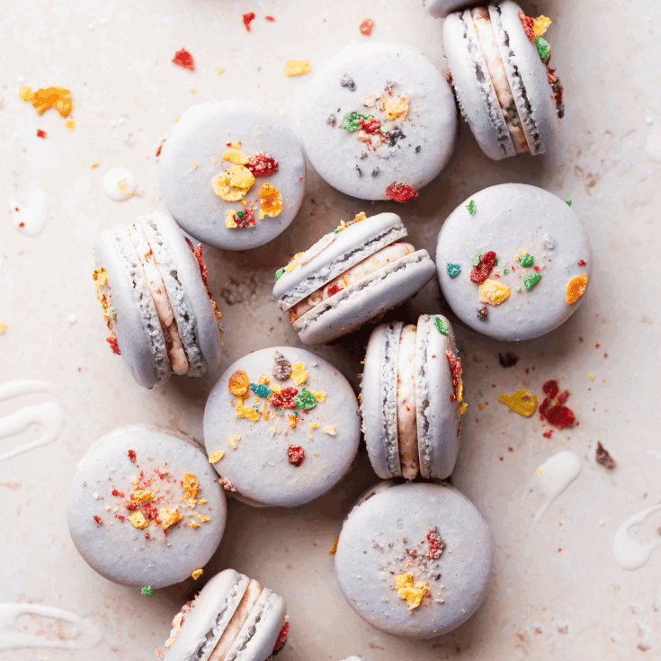 Several fruity pebbles macarons laying flat and some laying on their side on a white counter top with scattered pieces of fruity pebbles cereal.