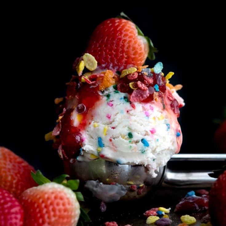 A metallic bowl with a large scoop of fruity pebbles ice cream on a dark counter top garnished with a whole strawberry and more fruity pebbles.