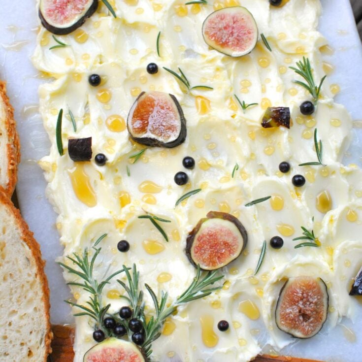 A white serving board with butter spread out, sliced figs, sprigs of rosemary and drizzled with honey on a white counter.