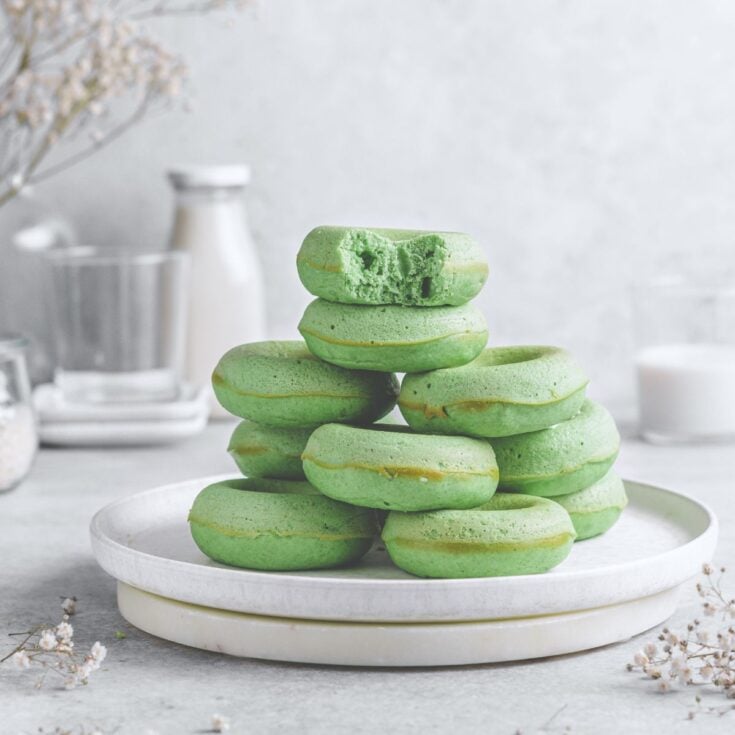 A large pile of several matcha donuts on a white plate on a white counter top with the top donut missing a bite.