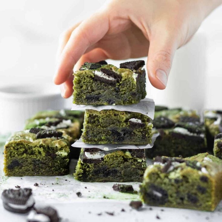 Several matcha Oreo brownies resting on white parchment paper with three brownies stacked on top of each other and a hand reaching for the top one.