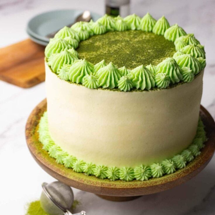 A whole frosted matcha cake dusted with matcha powder resting on a wooden cake stand.