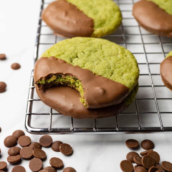 Four matcha green tea cookies with half dipped in chocolate resting on a wire rack with one of the cookies missing a bite and several chocolate chips scattered around on a white counter top.