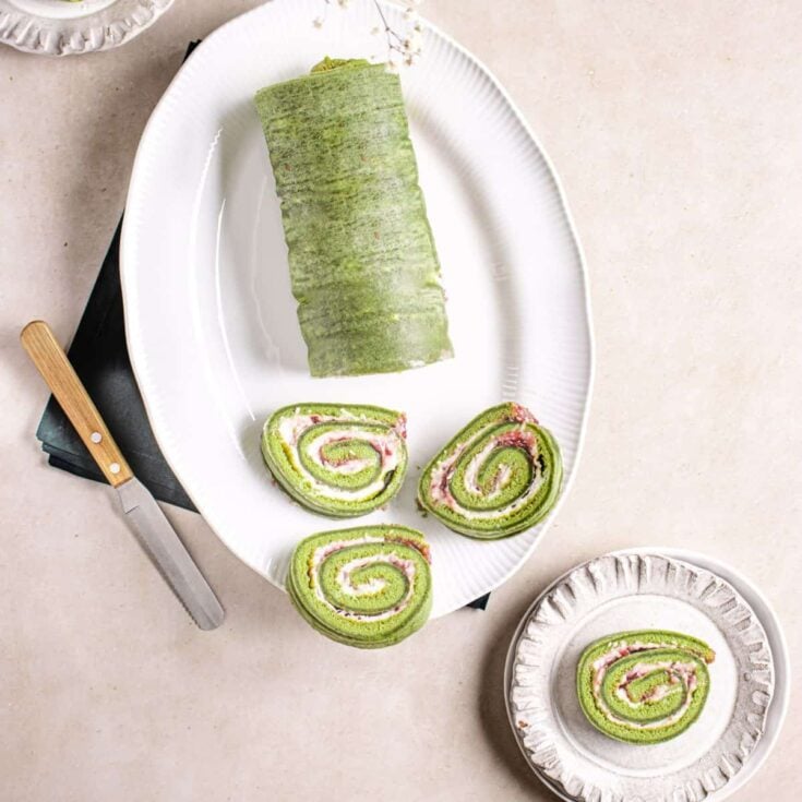 A large matcha roll cake on a white serving platter with three slices laying flat and one slice resting flat on a white serving plate.