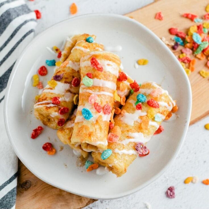 Four pieces of rolled fruity pebbles French toast on a white plate drizzled with white icing and garnished with pieces of fruity pebbles.