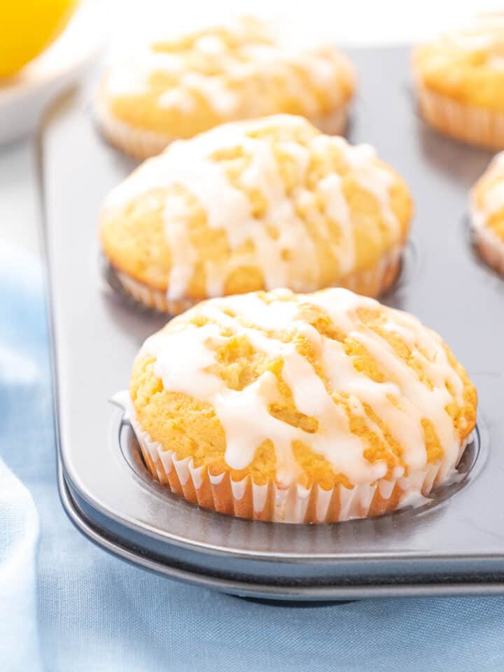 A six cup muffin tin filled with glazed lemon muffins on a white counter top with a light blue napkin.