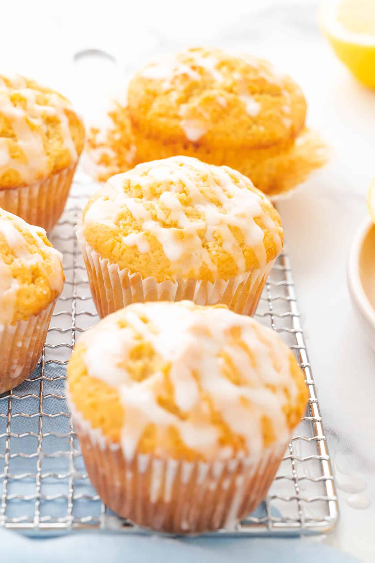 Five lemon glazed muffins resting on a wire rack on a white counter top with a light blue napkin.