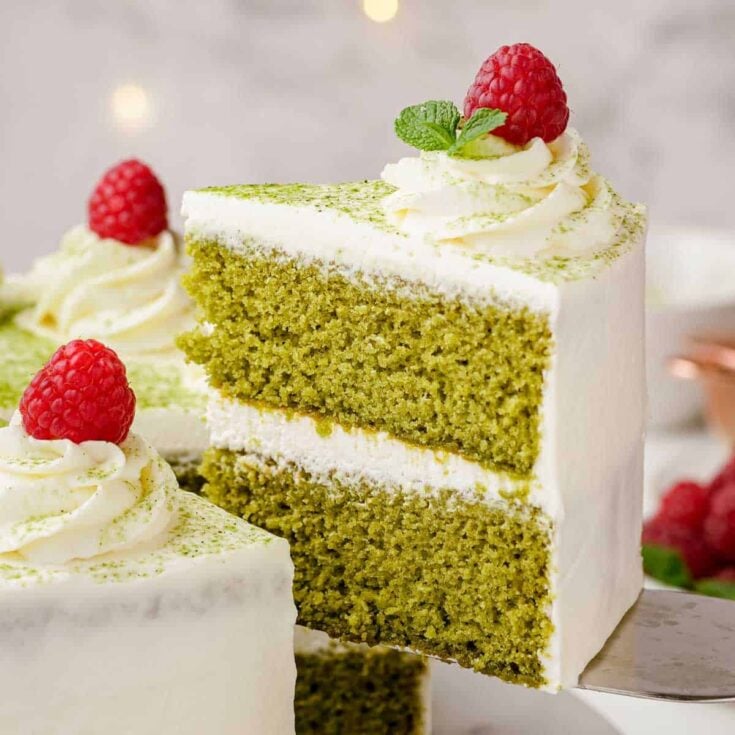 A large slice of matcha cake with white frosting being lifted away from a whole matcha cake.