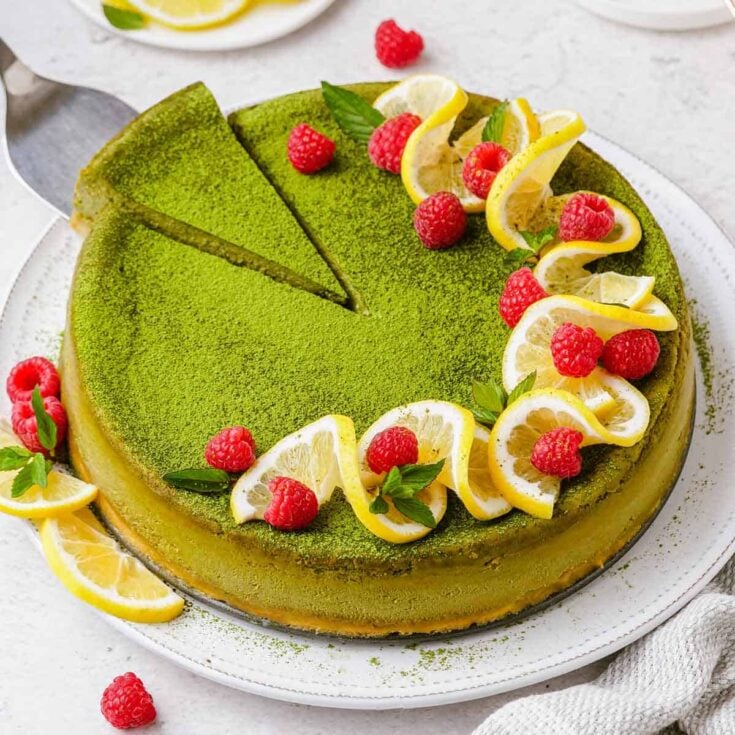 A large matcha cheesecake on a white serving platter garnished with fresh raspberries and lemon slices with one piece being removed with a serving knife.