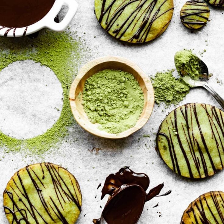 Three green matcha cookies drizzled with chocolate sauce on white parchment paper with a small wooden bowl of matcha powder.