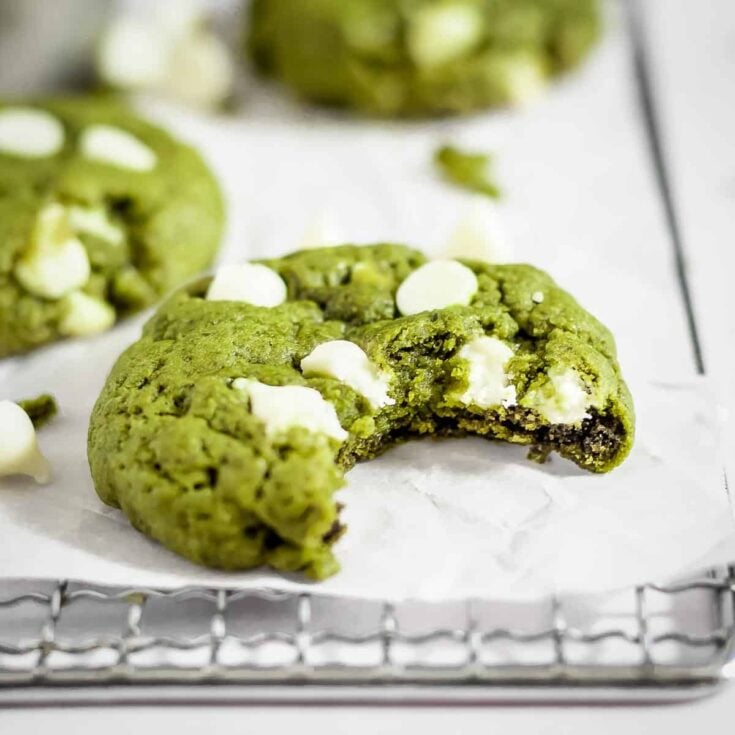 One large white chocolate chip matcha cookie with a bite missing resting on parchment paper on top of a wire rack on a white counter top.