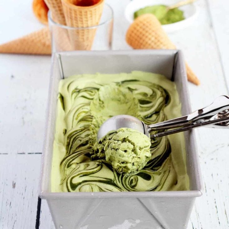 A large rectangular metal pan filled with matcha ice cream with an ice cream scoop and empty ice cream cones scattered about on a white wooden counter top.