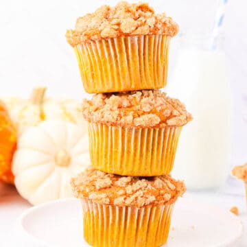 Three pumpkin muffins with cake mix stacked on top of each other on a white plate with three orange and white pumpkins and a glass of milk with a blue and white striped straw.