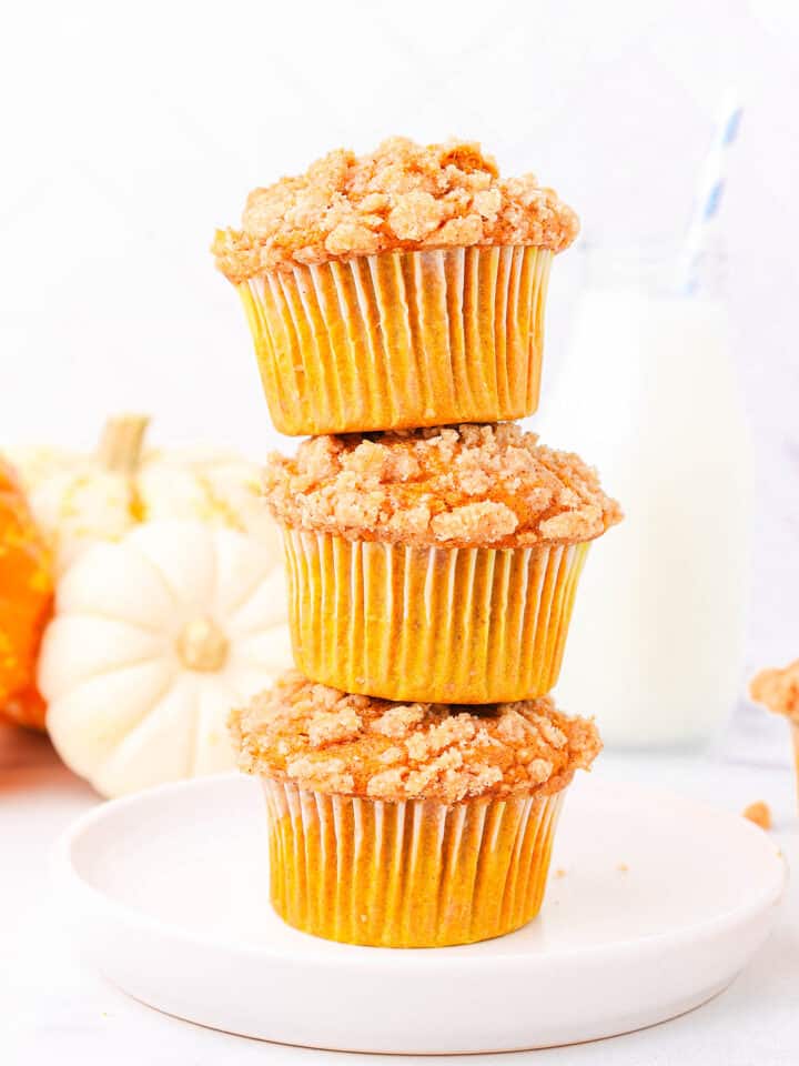 Three pumpkin muffins with cake mix stacked on top of each other on a white plate with three orange and white pumpkins and a glass of milk with a blue and white striped straw.