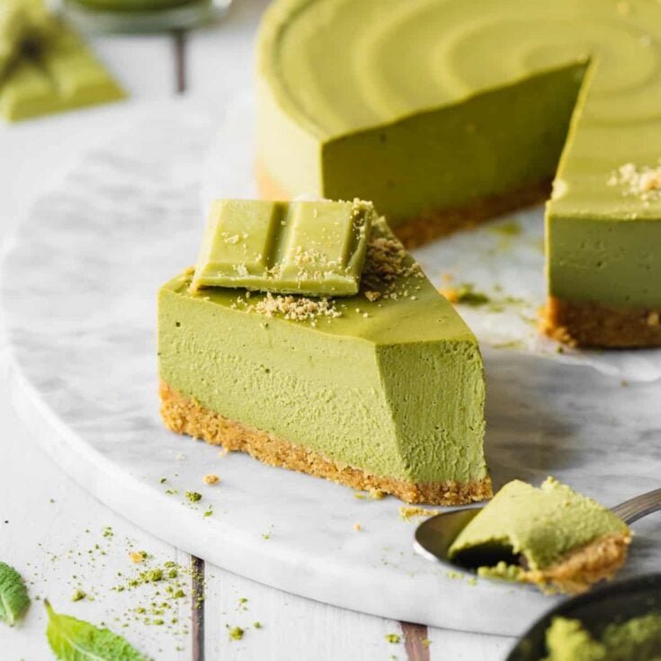 A large green matcha cheesecake with one slice taken out and placed on a white serving board.