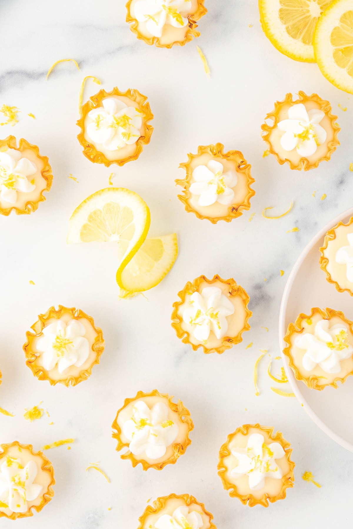 Several mini lemon tarts topped with whipped cream and fresh lemon zest on a white counter top with slices of lemon.
