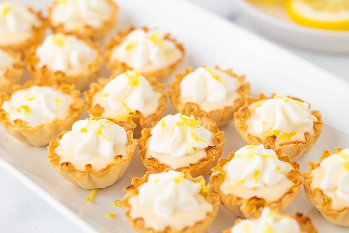 Several mini lemon curd tarts with phyllo shells topped with whipped cream and lemon zest on a white serving platter.