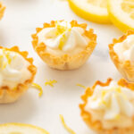 Five mini lemon tartlets on a white counter top with lemon zest sprinkled around and three lemon slices.
