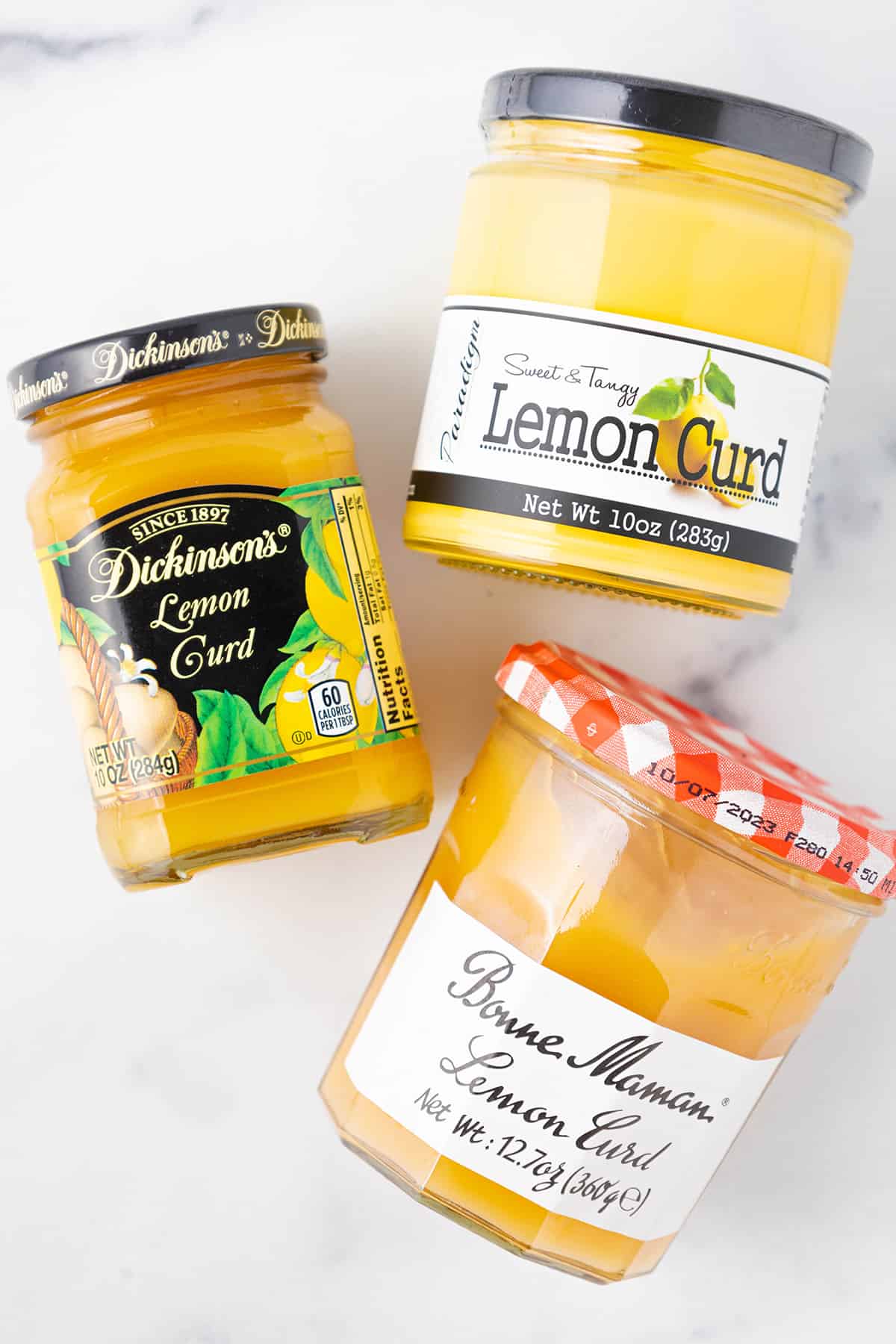 Three different brands of jarred store bought lemon curd including paradigm lemon curd, dickinsons lemon curd, and bonne maman lemon curd on a white counter top.