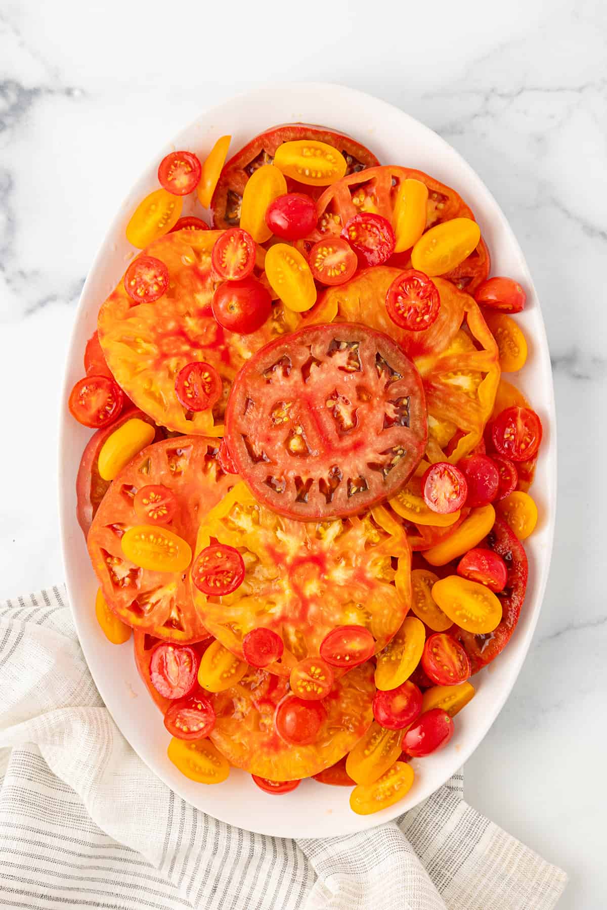 A large white platter with several slices of heirloom tomatoes, and several orange and red cherry tomatoes with a grey and white striped napkin on a white counter top.