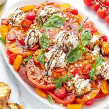 A large white serving platter with caprese salad with burrata on a white counter top with three pieces of crostini and several red cherry tomatoes scattered about.