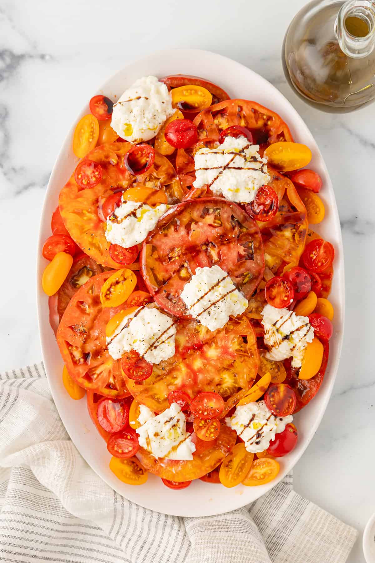 A large white serving platter with several sliced heirloom tomatoes, several sliced red and orange cherry tomatoes, and several pieces of torn burrata cheese drizzled with balsamic glaze and olive oil on a white counter top.