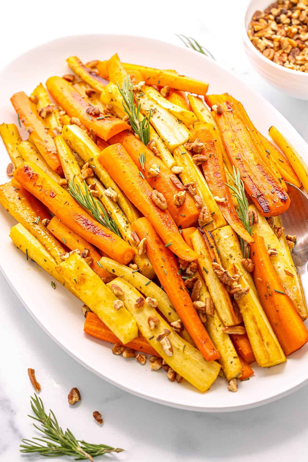 A large white platter with carrots and parsnips topped with fresh rosemary and chopped pecans on a white counter top.