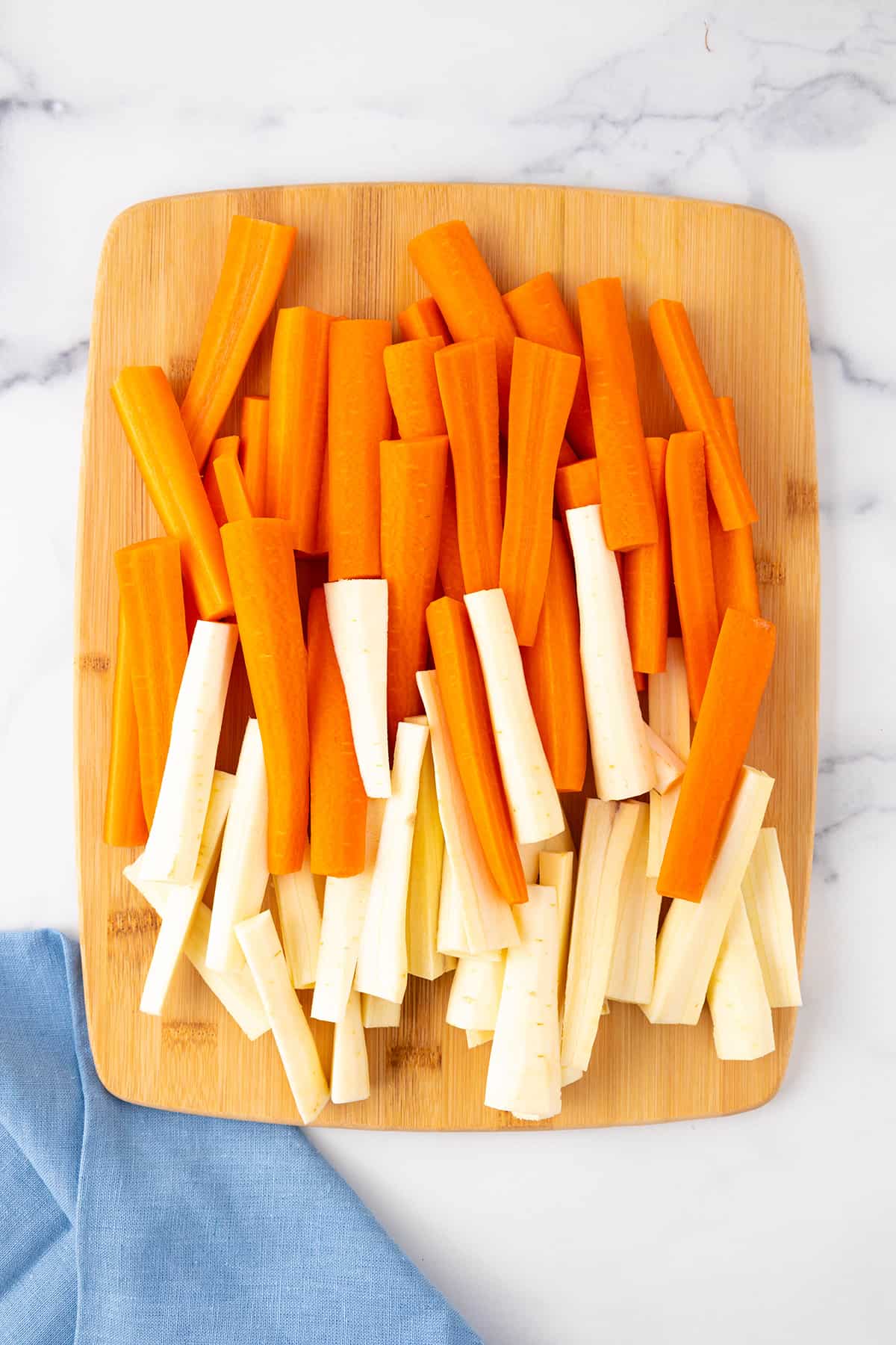 A wooden cutting board with carrots and parsnips that have been cut into spears with a blue napkin laying nearby.