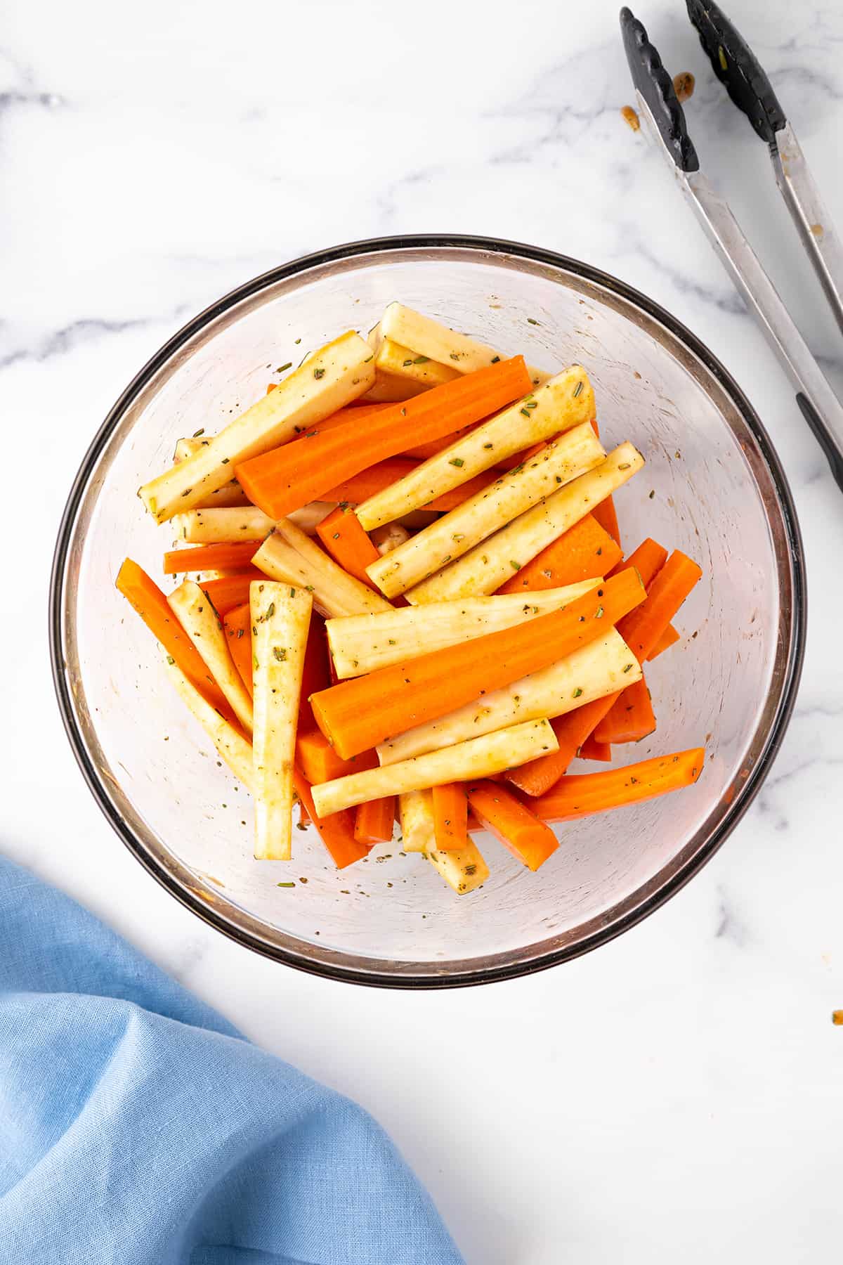 A large glass mixing bowl with spears of carrots and parsnips tossed in, honey, balsamic vinegar and rosemary on a white counter top.