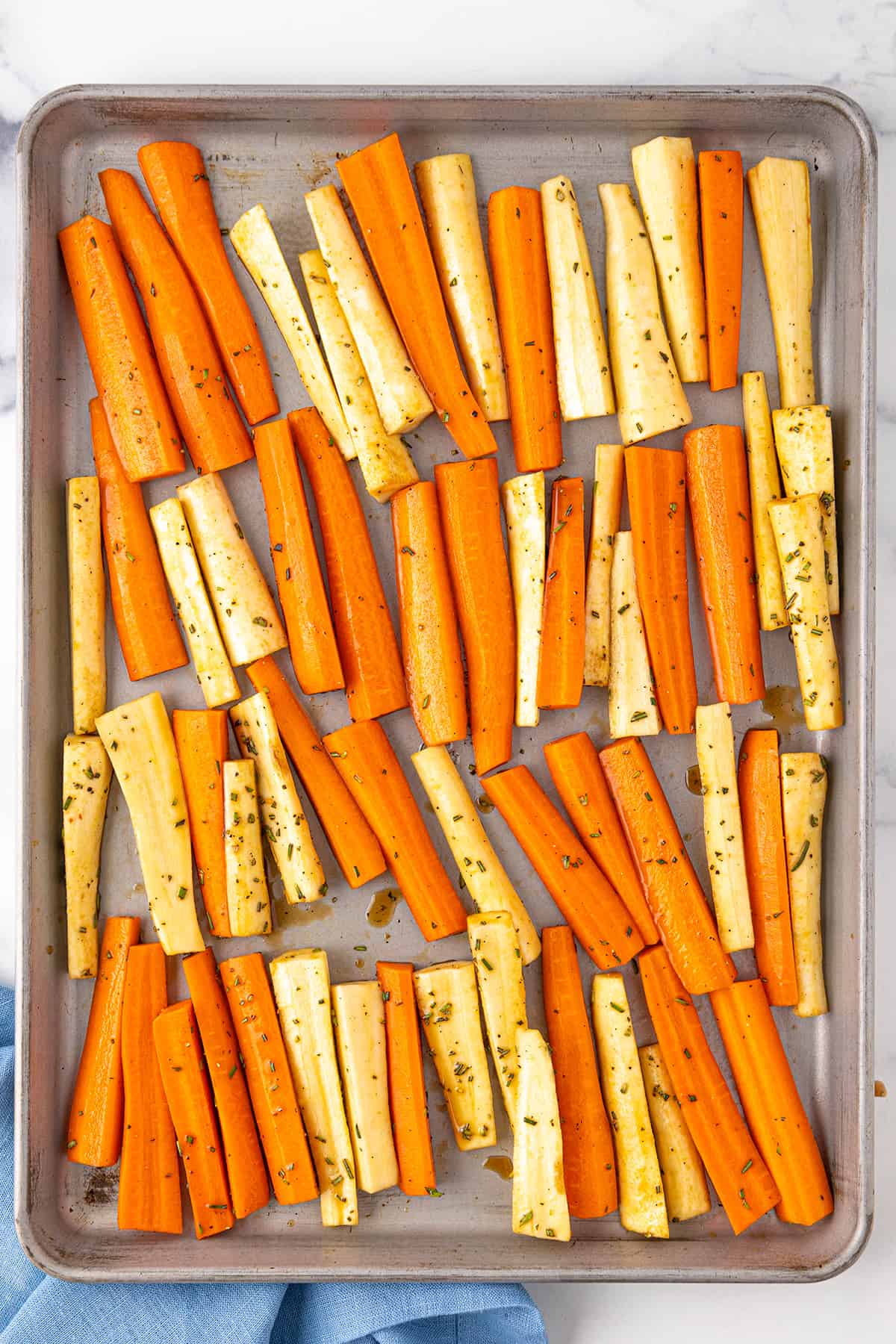 A large silver sheet pan with carrots and parsnips that have been cut into spears and tossed with honey, balsamic vinegar and fresh rosemary.