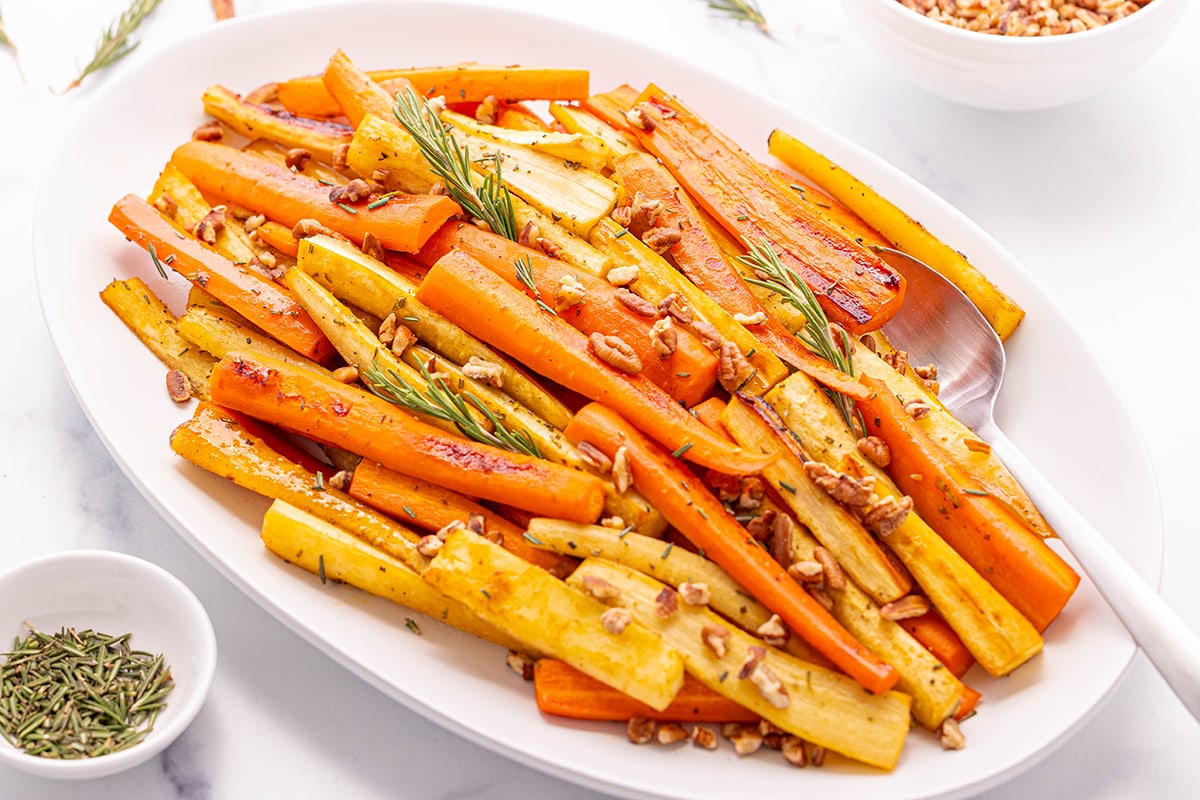 A large white oval platter with honey roasted carrots and parsnips with a small white bowl of chopped pecans and a small white bowl of chopped fresh rosemary on a white counter top.