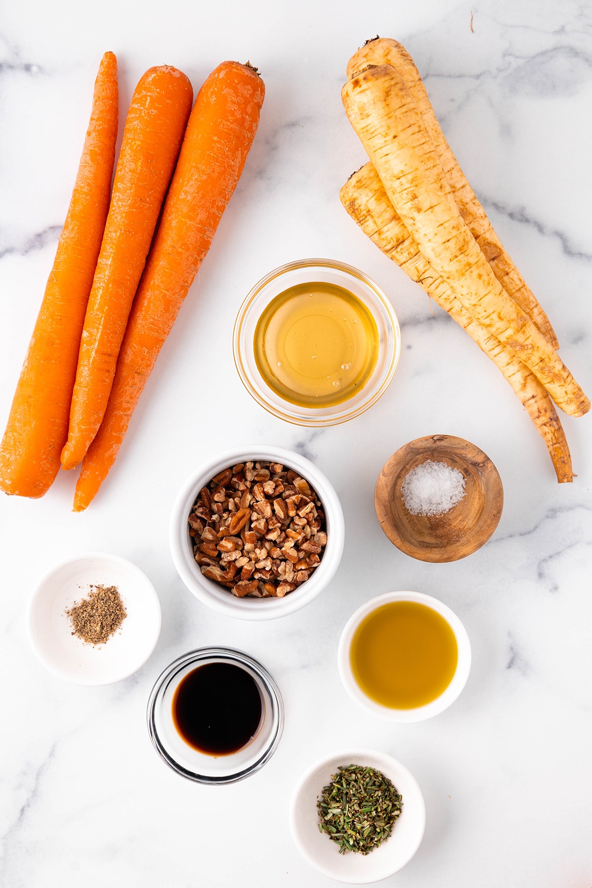 Three large carrots, three large parsnips, and small individual bowls with honey, chopped pecans, olive oil, balsamic vinegar, chopped rosemary, salt, and pepper on a white counter top.