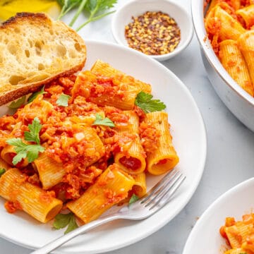 A large white bowl and a stainless skillet filled with rigatoni amatriciana on a white counter top with two pieces of toasted bread, some fresh parsley sprigs and a small bowl of red pepper flakes.