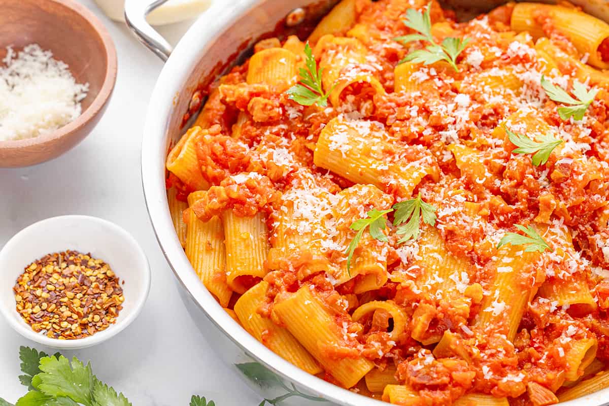 A large stainless steel skillet with rigatoni all'amatriciana on a white counter top with a small wooden bowl of shredded pecorino romano cheese and a small white bowl with red pepper flakes.