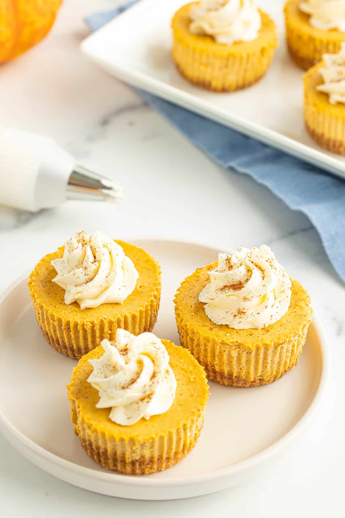 Three miniature pumpkin cheesecakes topped with fresh homemade whipped cream on a white dessert plate and three more cheesecakes on a serving platter with a piping bag of whipped cream on a white counter top.