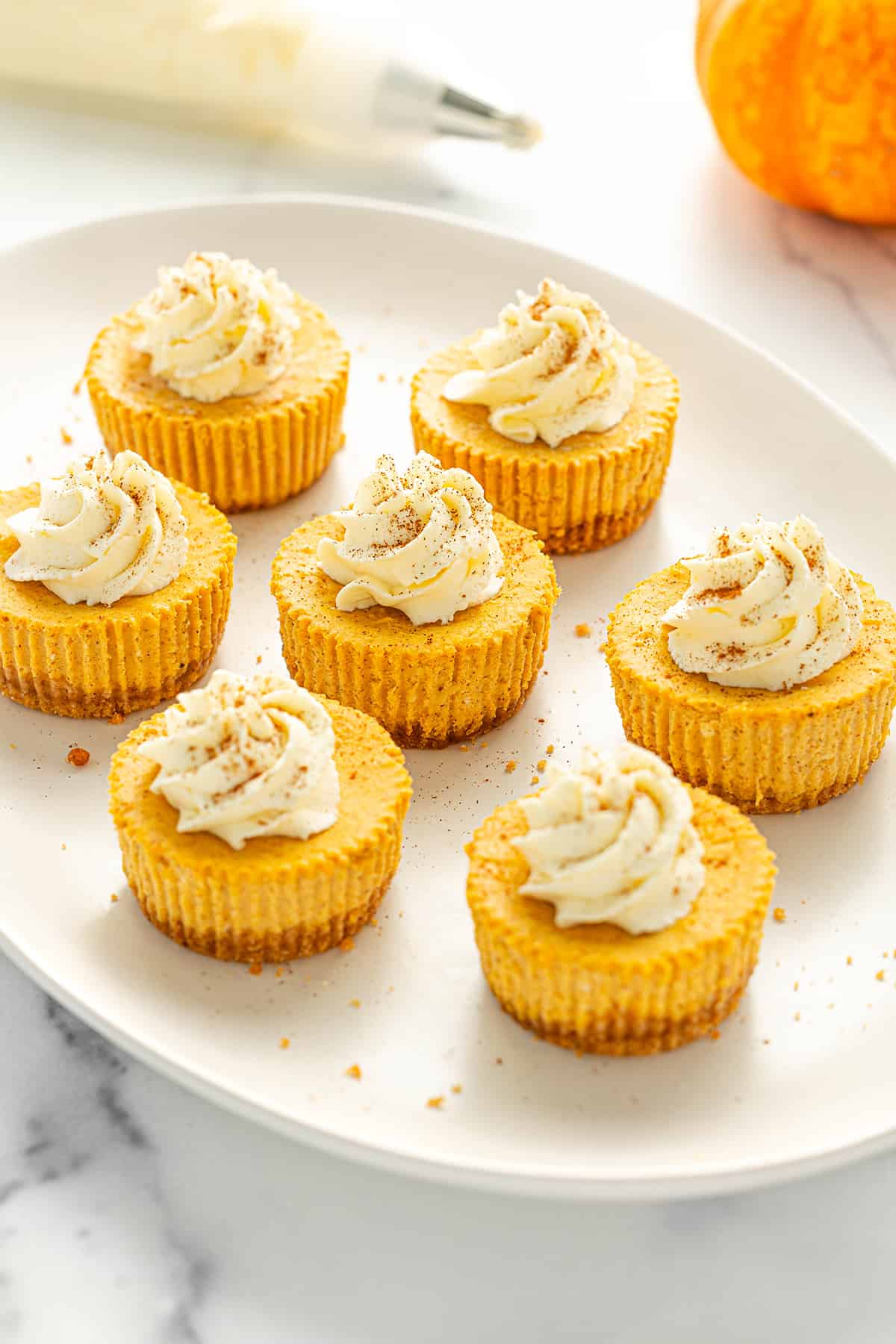 Seven mini pumpkin cheesecakes topped with whipped cream and a sprinkle of cinnamon on an oval serving platter with an orange pumpkin and a piping bag filled with whipped cream on a white counter top.