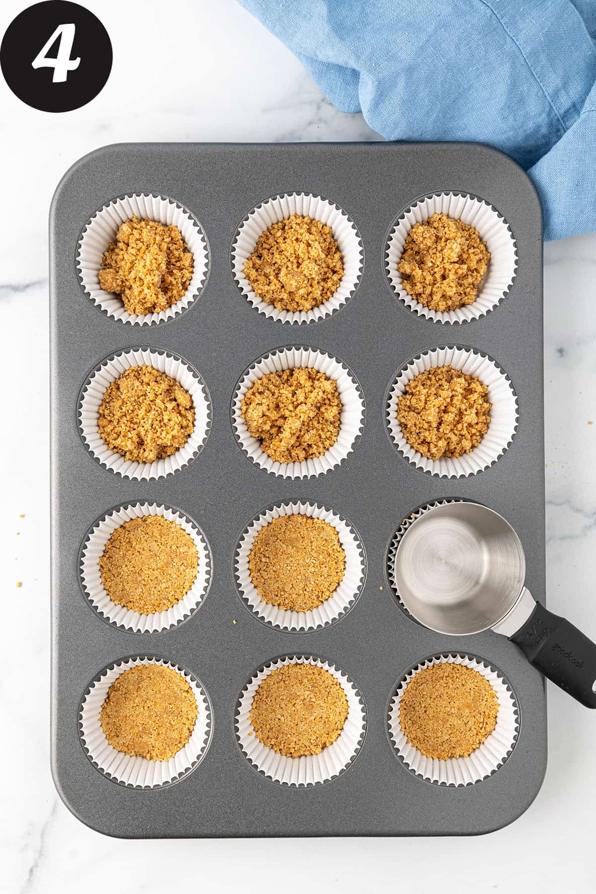 A regular sized muffin pan filled with paper cupcake liners, filled with a graham cracker butter mixture and a flat bottomed measuring cup pressing the crumbs down flat to form a crust.