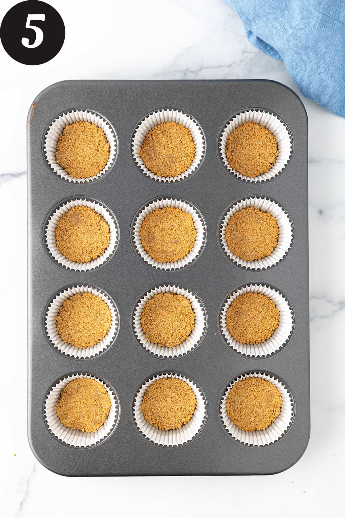 A twelve cup muffin pan filled with white paper liners each with a backed graham cracker crust on a white counter.