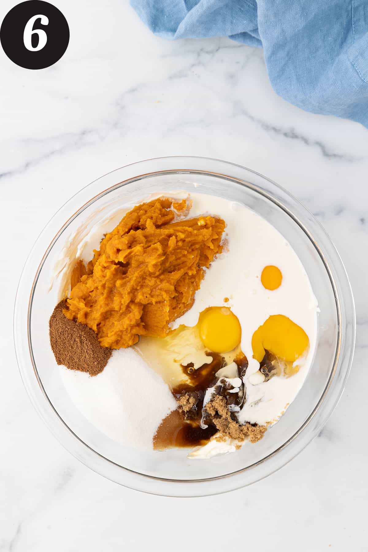 A large glass bowl on a white counter top filled with cream cheese, pumpkin puree, pumpkin pie spice, white sugar, brown sugar, three cracked eggs, vanilla extract and heavy cream.