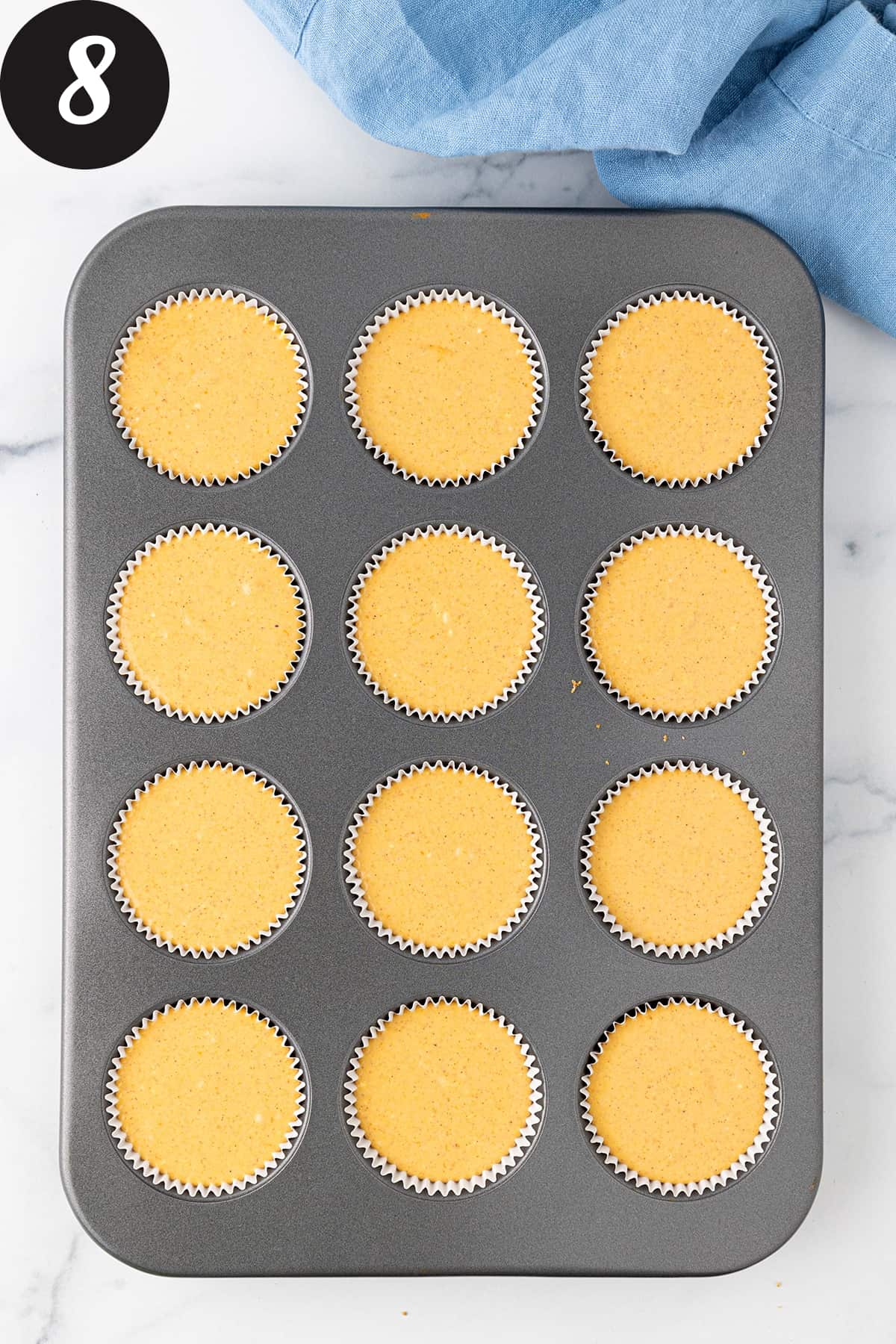 A twelve cup muffin pan lined with paper muffin liners filled with pumpkin cheesecake batter on a white counter top.