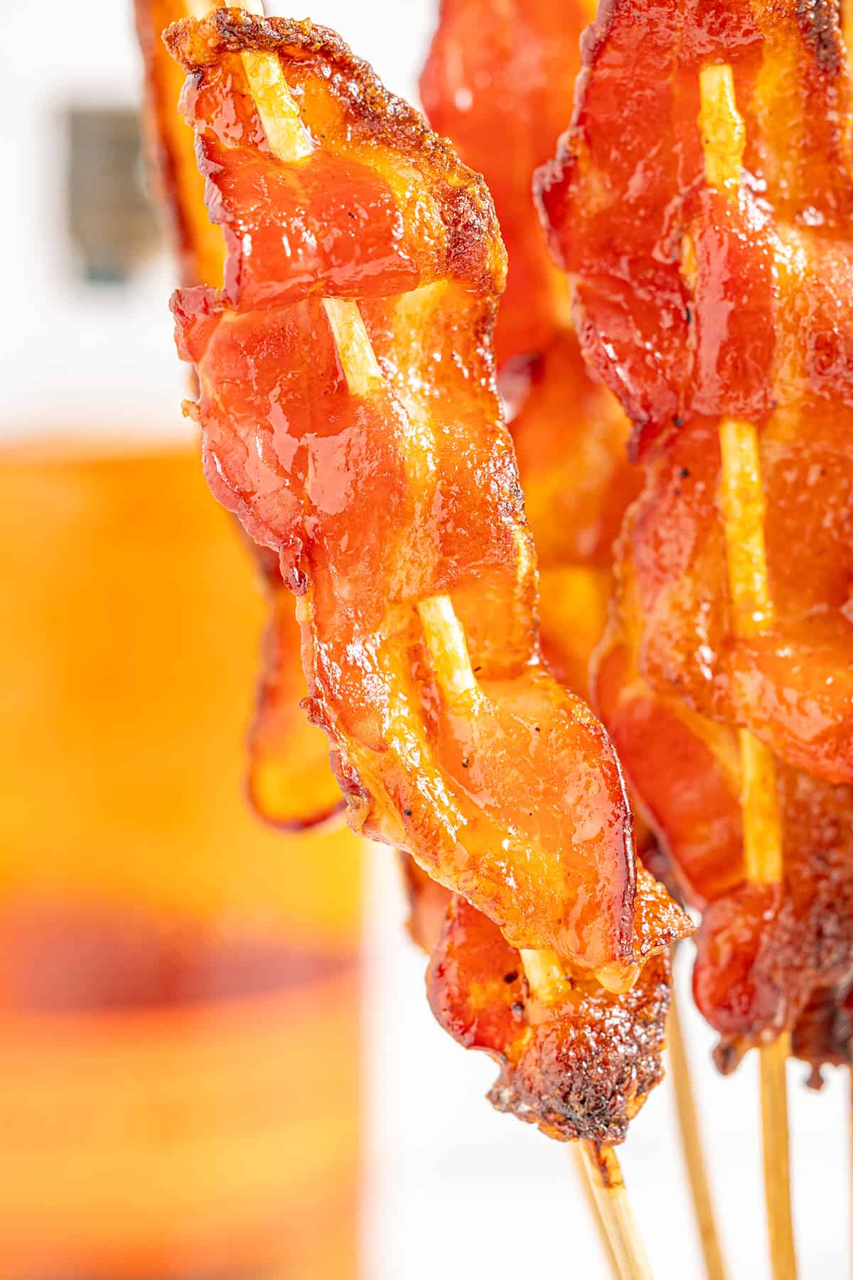 Candied bacon on a stick standing upright on a counter with a bottle of bourbon in the background.