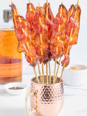 Several candied bacon skewers standing upright in a copper mug on a white counter top with a bottle of bourbon and a white ramekin of brown sugar.