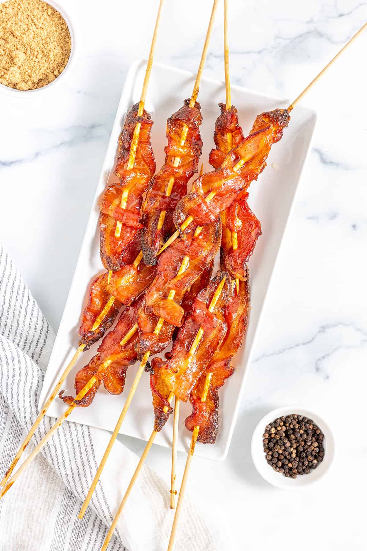 Several candied bacon skewers on a white platter on a white counter top with a striped napkin and a ramekin of brown sugar and a small bowl of whole peppercorns.
