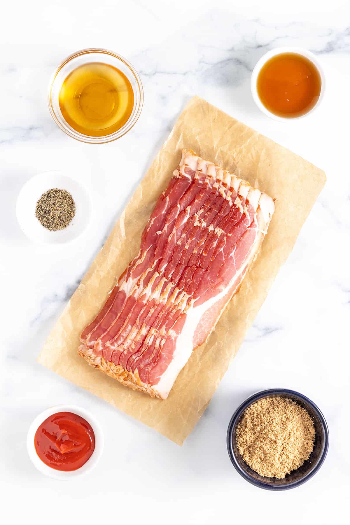 Several slices of raw bacon on a piece of parchment paper, light brown sugar, ketchup, maple syrup, bourbon, and ground black pepper on a white counter top.