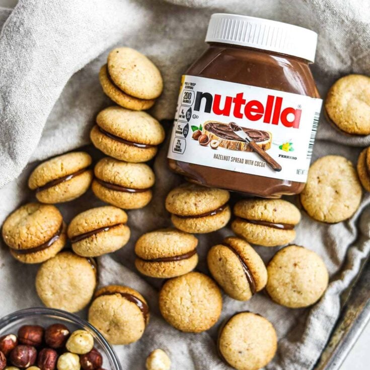 Several baci di dama cookies on a gray napkin with a small glass bowl of hazelnuts and a jar of Nutella.