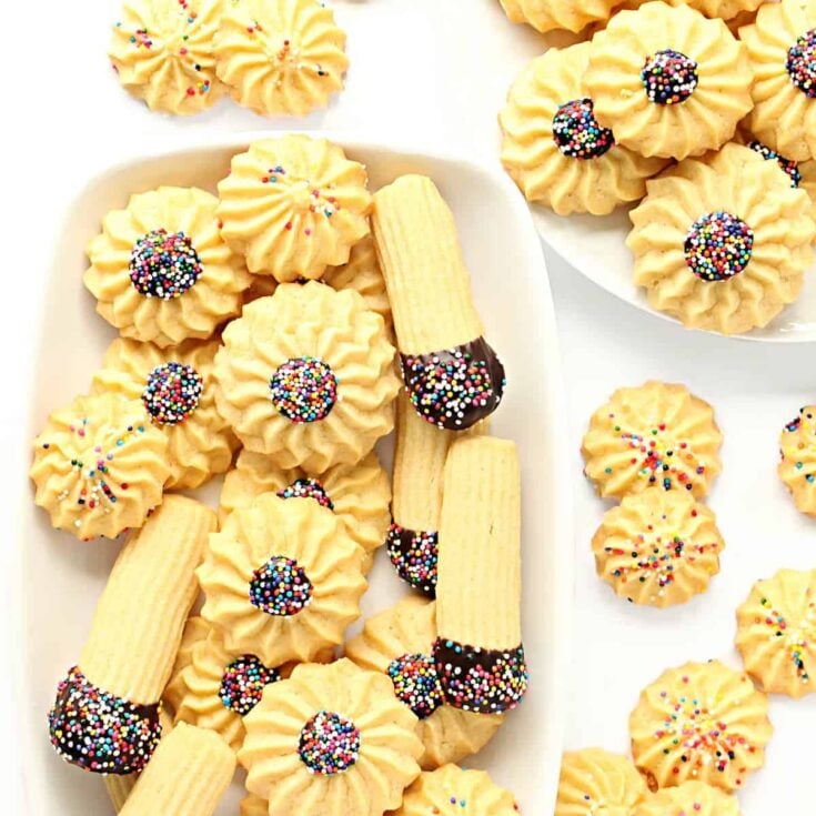 Several Italian butter cookies some dipped in chocolate with sprinkles on a white serving platter on a white counter top.