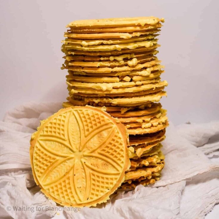 A large stack of several anise pizzelle and one anise pizzelle cookie leaning up against the stack on a white napkin.