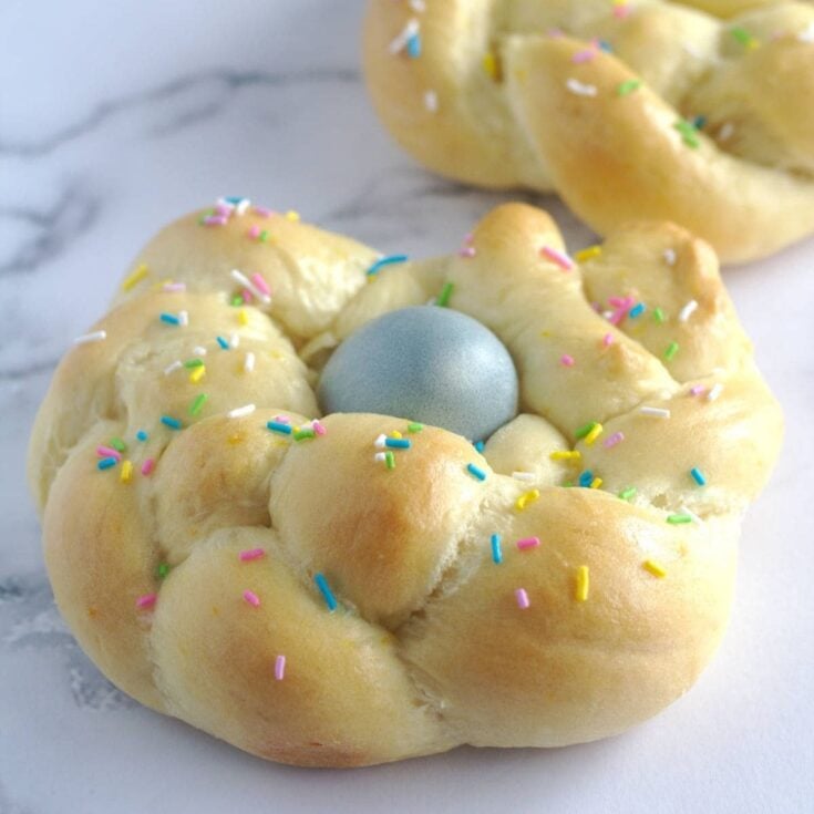 Italian easter bread in the shape of a braid with a blue egg in the middle topped with multicolored pastel sprinkles on a white counter top.