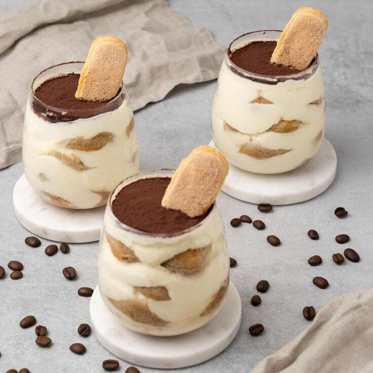 Three mini tiramisu cups on a light counter top with several espresso beans scattered about.